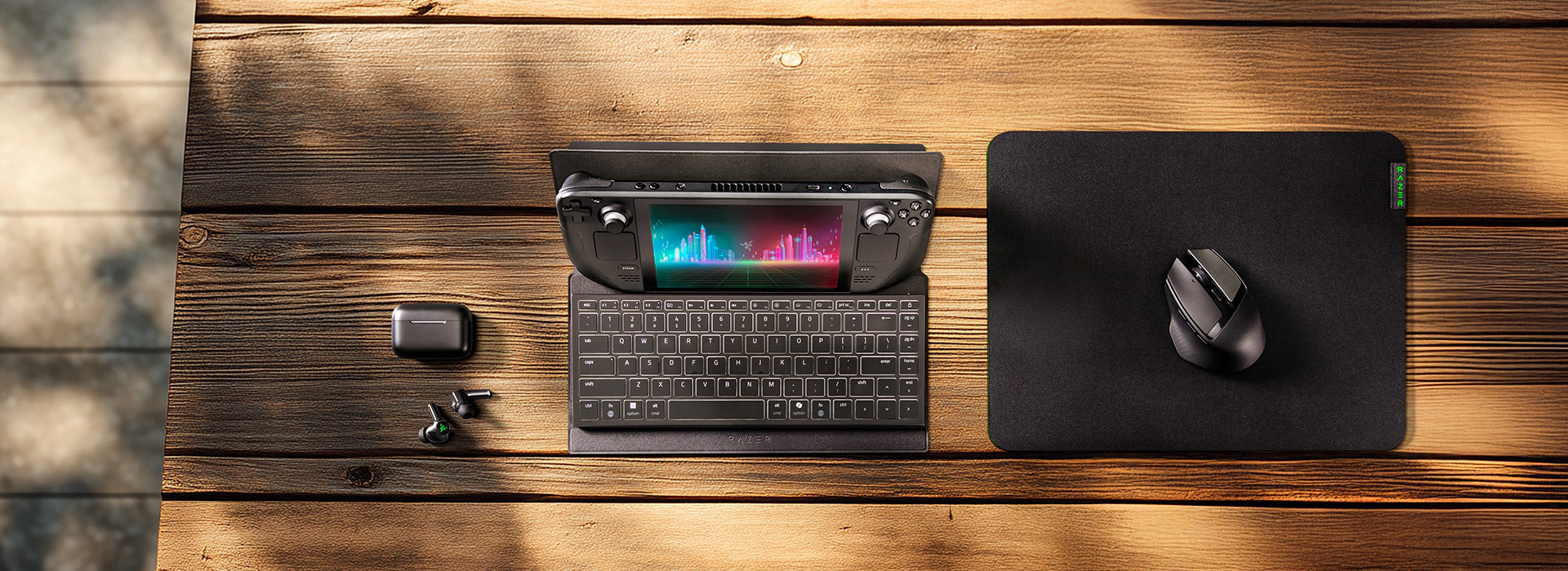 Handheld gaming device docked in a keyboard travel case, next to wireless earbuds and a mouse on a mousepad, arranged on a wooden table.