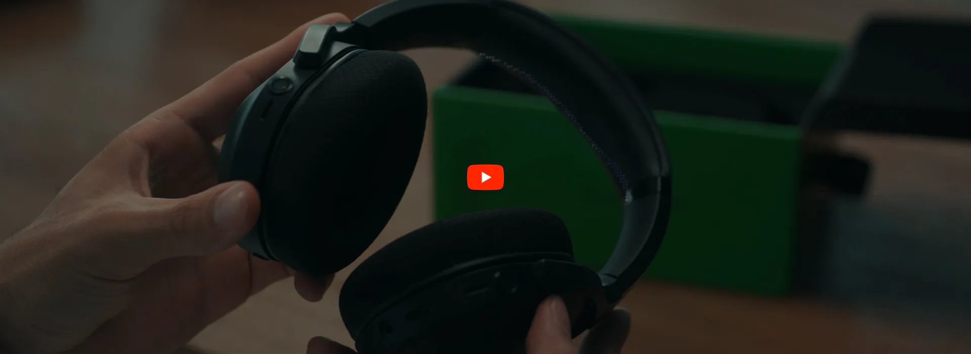 Hand holding black over-ear headphones, with a green box in the background, and a YouTube play button overlay.