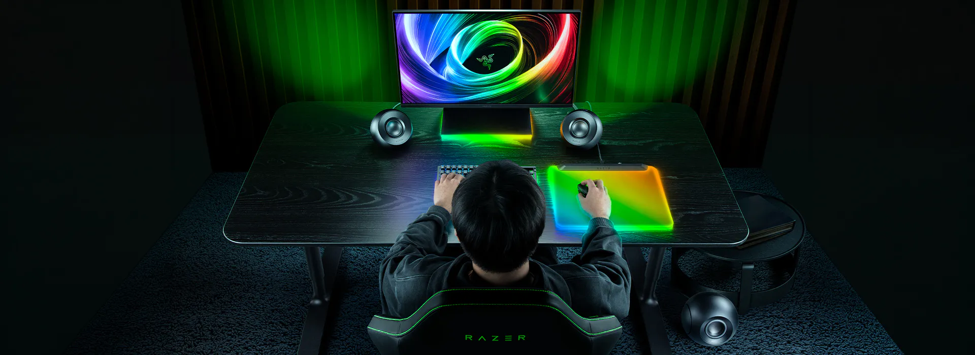 A person sits at a gaming desk with a Lenovo monitor, Razer speakers, RGB mousepad, and keyboard, surrounded by neon-green backlighting.