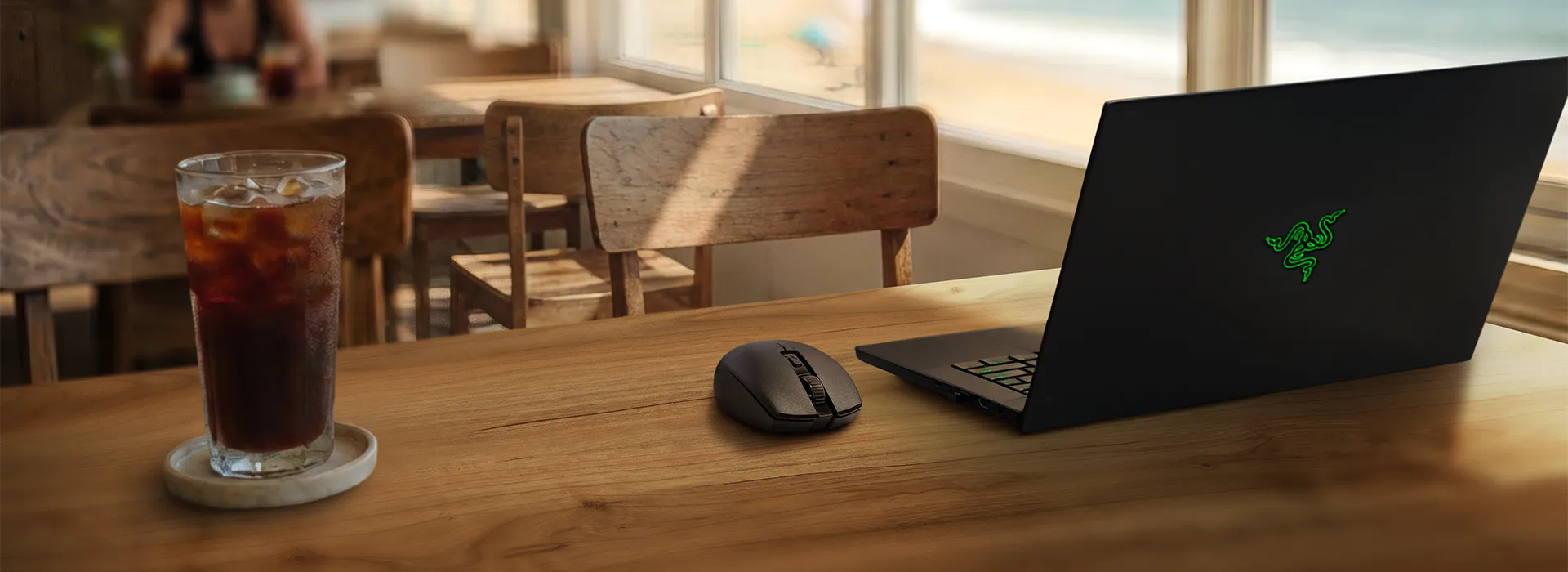 A wooden table with a glass of iced cola, a black gaming mouse, and a Razer laptop with a green snake logo, near a window.