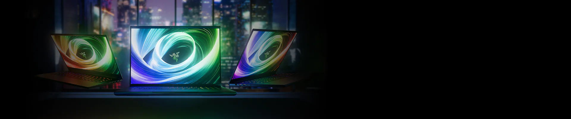 Three Razer laptops with colorful screens, open on a desk in a dark room with a city skyline view in the background.