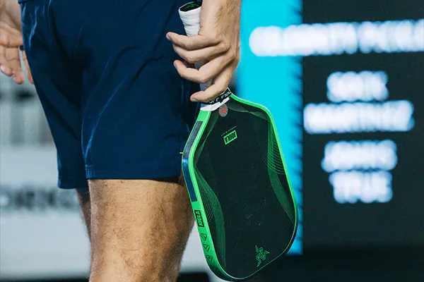 close-up of a person in blue shorts holding a green and black pickleball paddle by their side, standing near an led scoreboard with blurred text.
