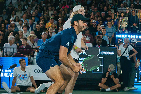 two players are playing pickleball in a crowded stadium, focused and poised ready to receive a serve. the crowd watches intently, creating an energetic atmosphere.
