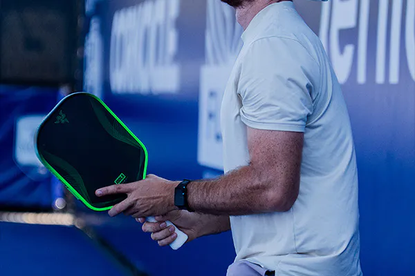 player's holding a green and black pickleball paddle, wearing a white shirt and black watch, standing near a blue wall with white text.