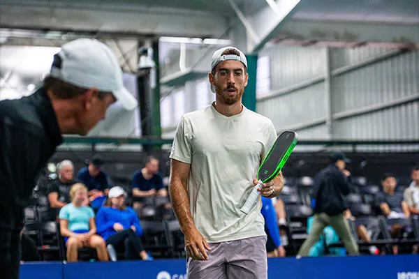 a player in a white t-shirt and cap holds a pickleball paddle on an indoor court, others sitting and walking in the background.