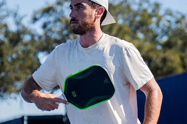 player is holding a green and black paddle, standing outdoors with trees in the background.