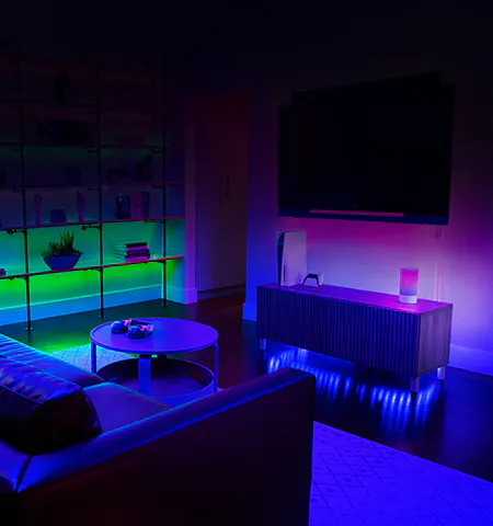 A modern living room illuminated with colorful LED lights, featuring a tv, bookshelf, plant, and a table with headphones.