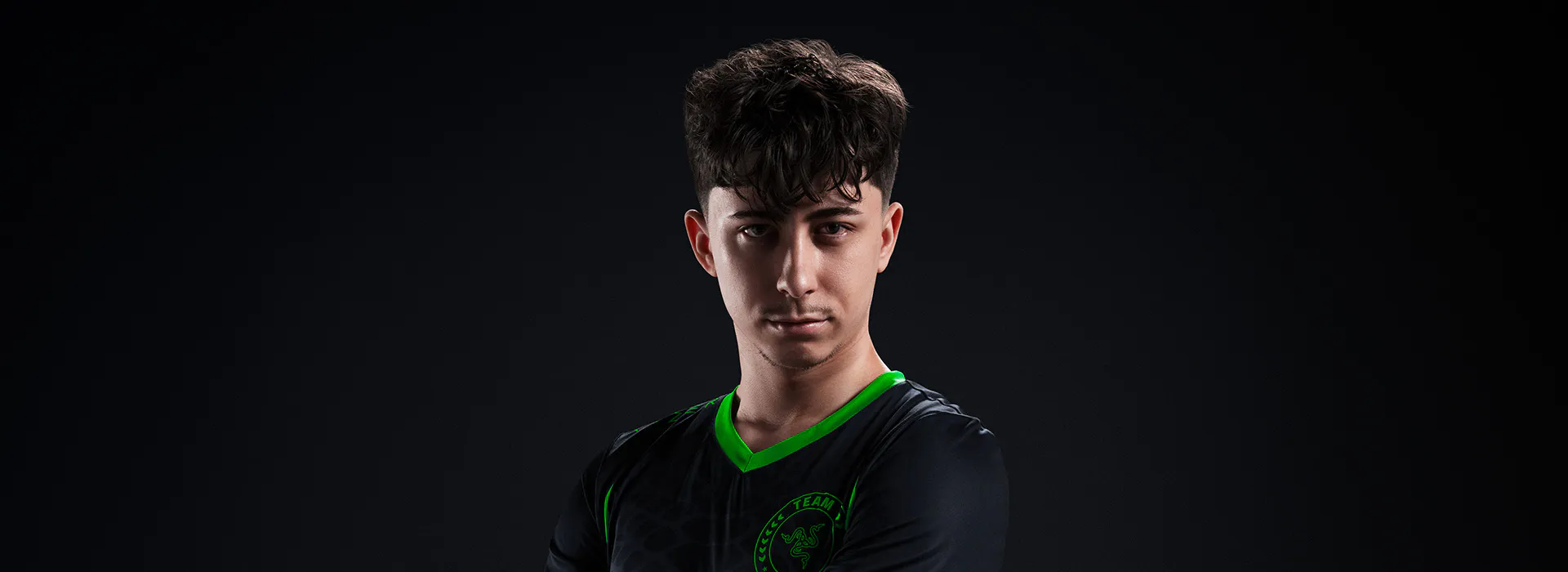 A young man with curly dark hair wearing a black jersey with green accents and a Razer Team logo, posing confidently against a black background.